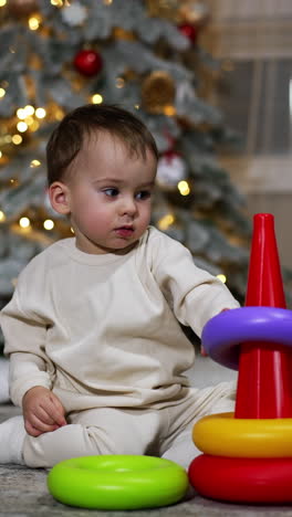 nice calm baby boy plays with a toy pyramid sitting on the floor. room decorated for christmas at backdrop. vertical video