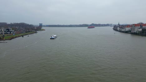 Aerial-Flying-Over-Oude-Maas-On-Cloudy-Overcast-Day-With-Boat-Sailing-Past-And-Container-Ship-Approaching-In-Distance-Background