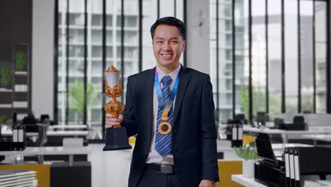asian business man in a suit and tie wears a gold medal and holds a gold trophy in his hand as the first winner smiling to camera in office