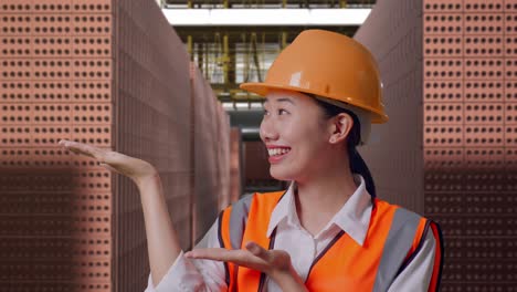 Close Up Of Asian Female Engineer With Safety Helmet Smiling And Pointing To Side While Standing With Red Brick Packed in Stacks Are Stored