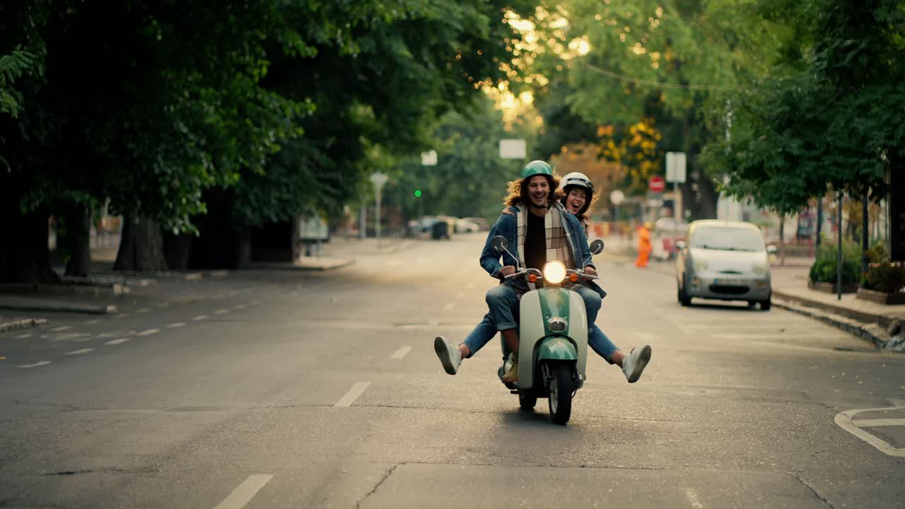 A happy guy with long curly hair in a denim jacket rides a moped on the road with his happy girlfriend and spreads his legs in different directions while riding on a busy morning city street