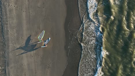 un par de windsurfistas saliendo del mar del norte caminando en una playa de arena con cometas al atardecer - vista aérea de arriba hacia abajo