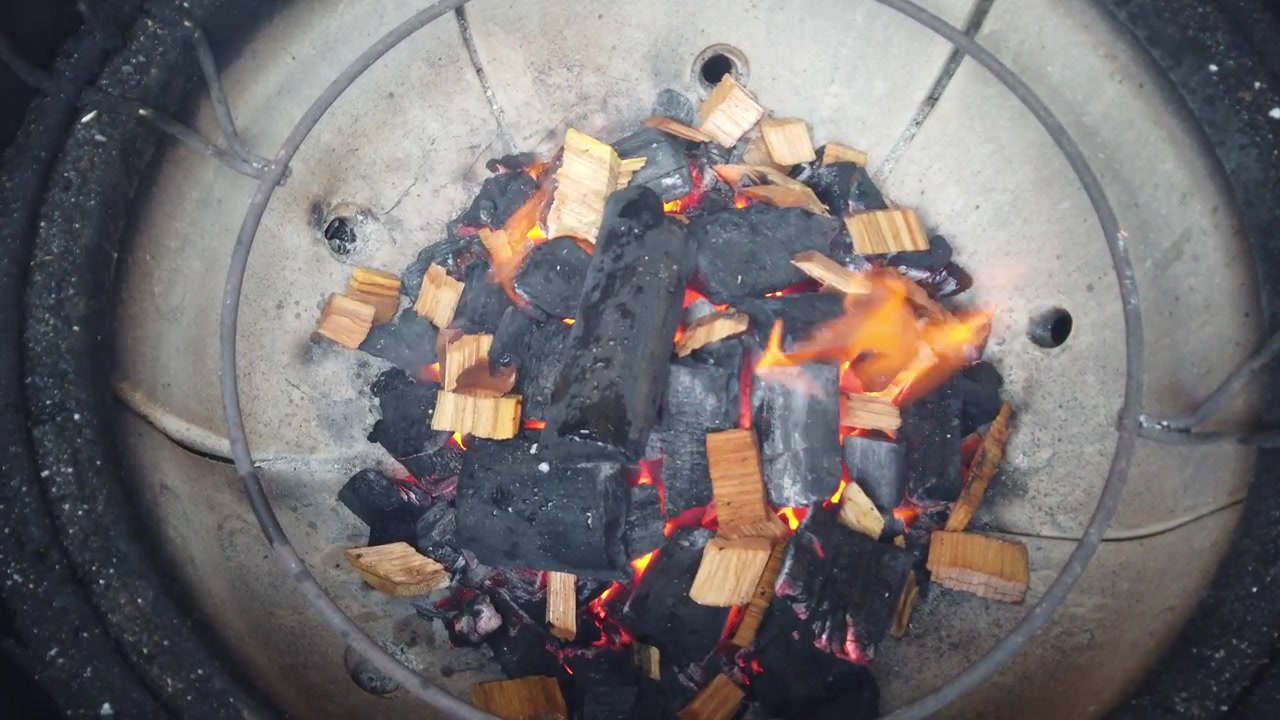 Pohled shora na hořící dřevěné uhlíkové brikety s kouřícími dřevěnými štěpkami uvnitř keramického grilu typu kamado, připravené k vaření a grilování.