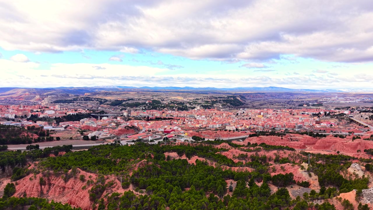 Ampia inquadratura panoramica della città di Teruel in Spagna, circondata da pinete e formazioni geologiche uniche di argilla rossa sotto un cielo nuvoloso.