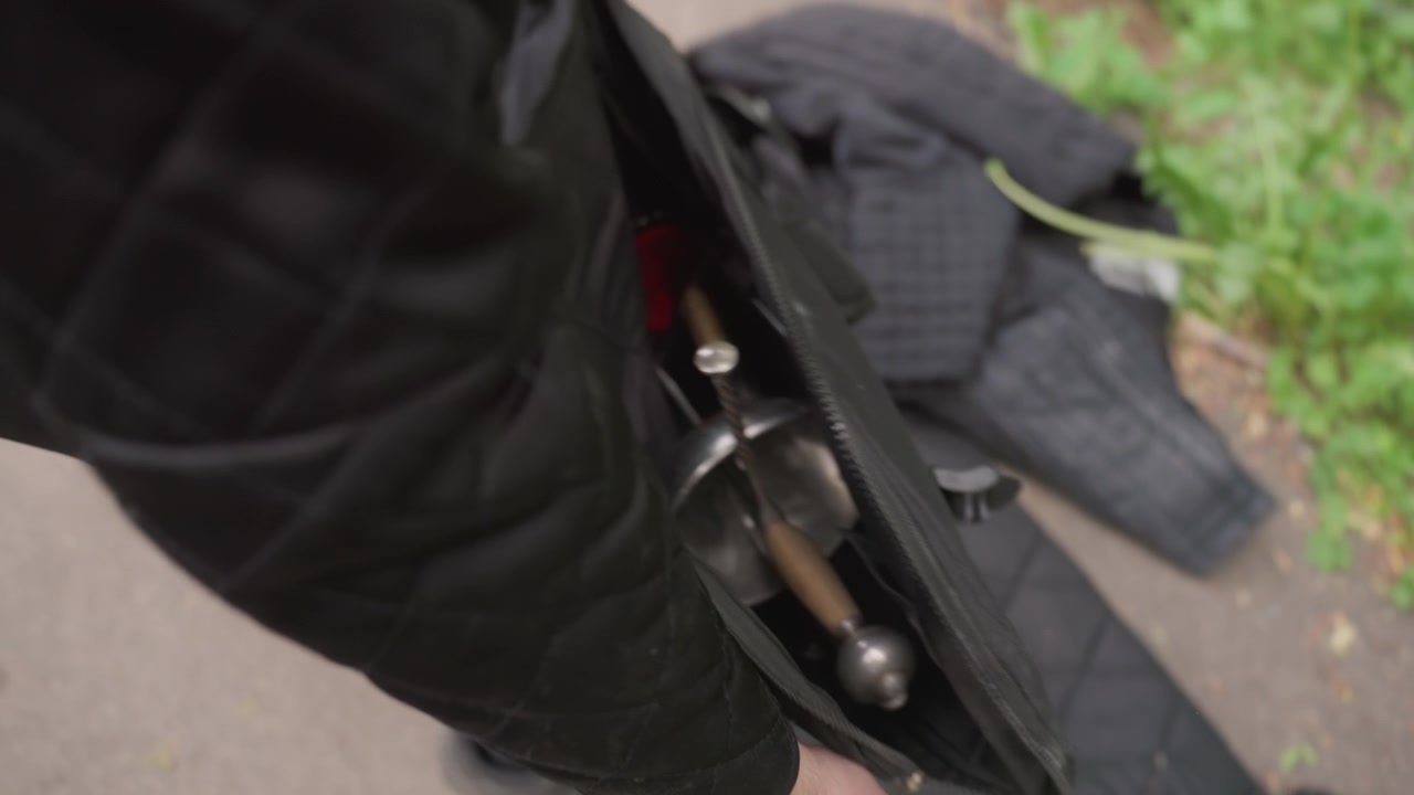 Close-up seseorang berbusana hitam memegang tas terbuka berisi peralatan anggar yang memperlihatkan pedang dan perlengkapan pelindung. Adegan menunjukkan tekstur detail bilah logam dengan gagang kayu