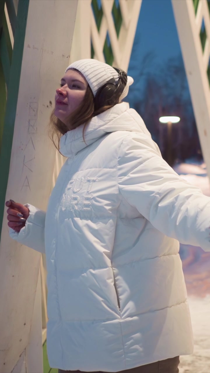 Woman in white puffer jacket and knit hat stands near outdoor wooden structure with graffiti including names written on it. Woman wears headphones or earmuffs and appears to be observing her