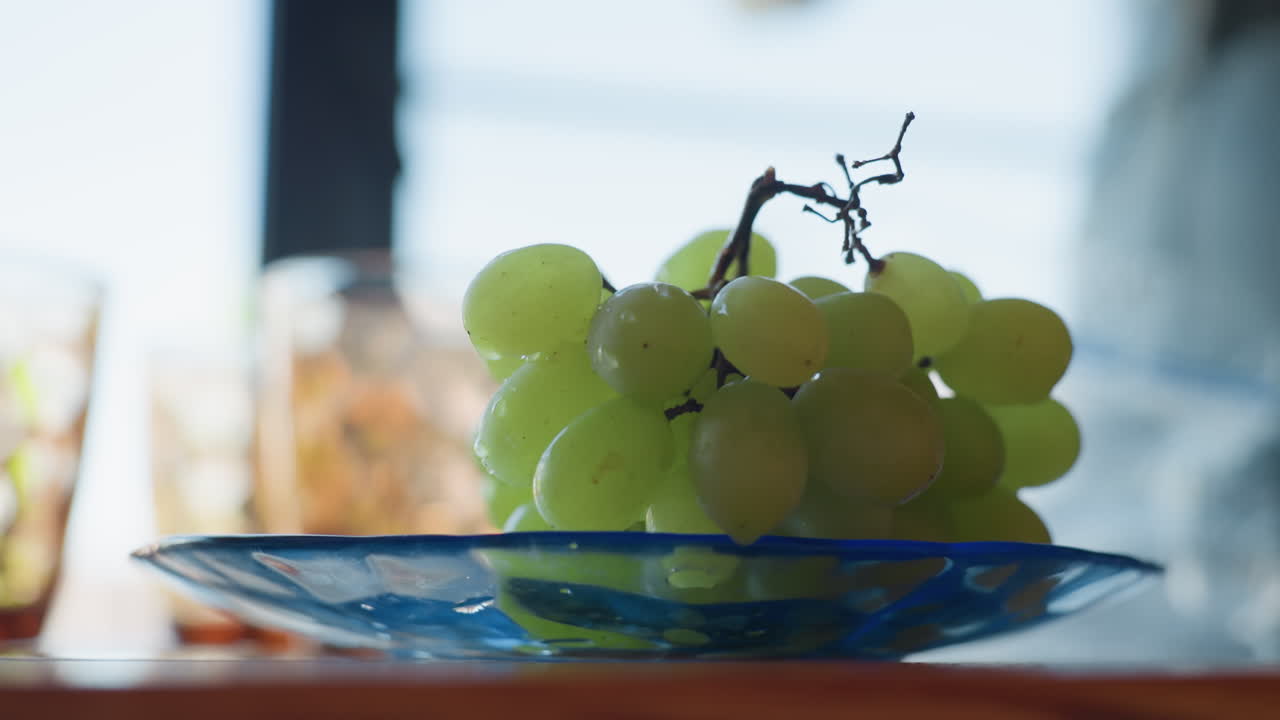 Cluster of fresh green grapes rests gently on transparent blue plate atop wooden table, daylight streaming softly from background, casting reflections