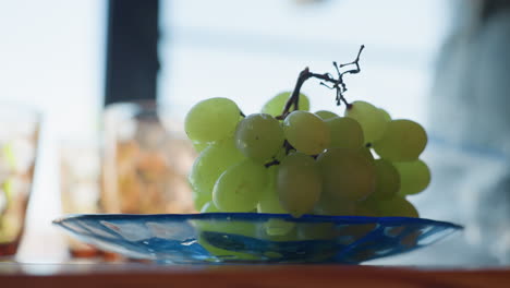 cluster of fresh green grapes rests gently on transparent blue plate atop wooden table, daylight streaming softly from background, casting reflections