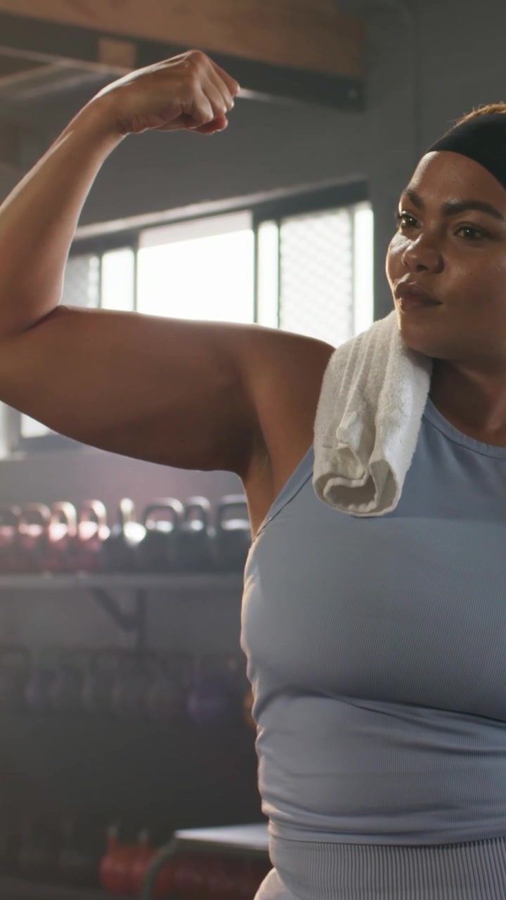 Vertical video: Adjusting towel, woman in blue top flexing left bicep at gym displaying strength