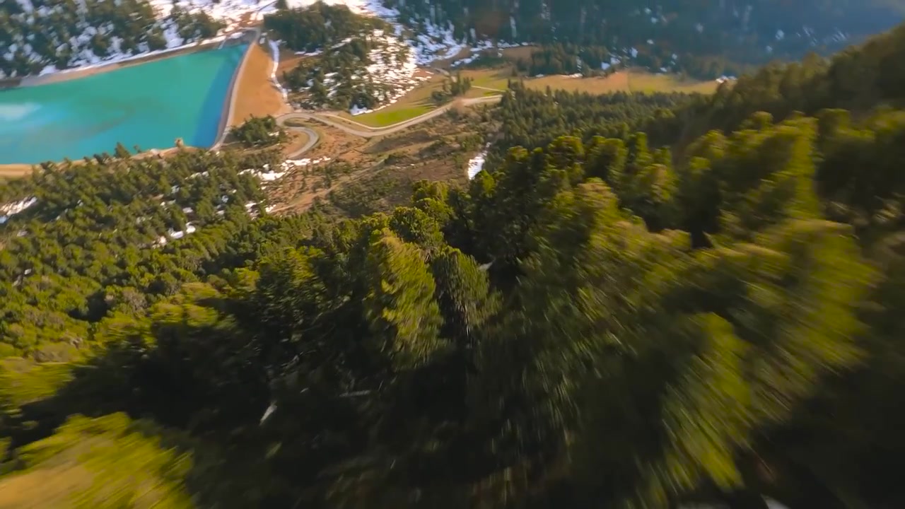 Drone FPV aéreo voando por uma crista ou encosta de montanha onde pinheiros altos crescem e são iluminados pela luz do sol. O drone chega ao sopé da montanha e um lago azul com neve é visível sob o sol.