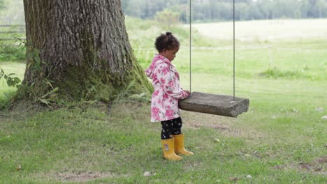 lonely adorable little african girl in raincoat waiting to play near swing, static, full shot, day