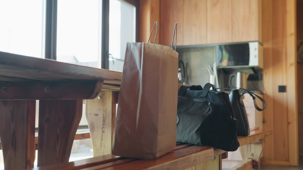 Three ladies stand around wooden bench inside warm cabin kitchen as they drop bags and prepare to leave, with soft sunlight streaming through large windows