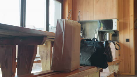 three ladies stand around wooden bench inside warm cabin kitchen as they drop bags and prepare to leave, with soft sunlight streaming through large windows