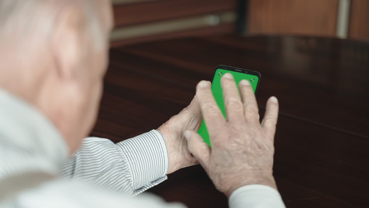 Over the shoulder shot of senior man sitting at the table in living room, holding modern smartphone with green screen in his wrinkled hands and doing video call in distance with family or friends