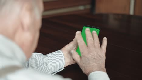 sobre el hombro de un anciano sentado en la mesa de la sala de estar, sosteniendo un teléfono inteligente moderno con pantalla verde en sus manos arrugadas y haciendo llamadas de video a distancia con familiares o amigos