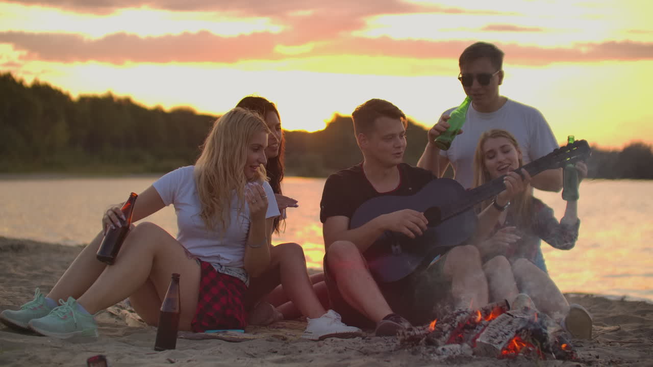 The five students play the guitar and sing songs around bonfire on the beach party. This is soulful summer evening with the best friends and beer.