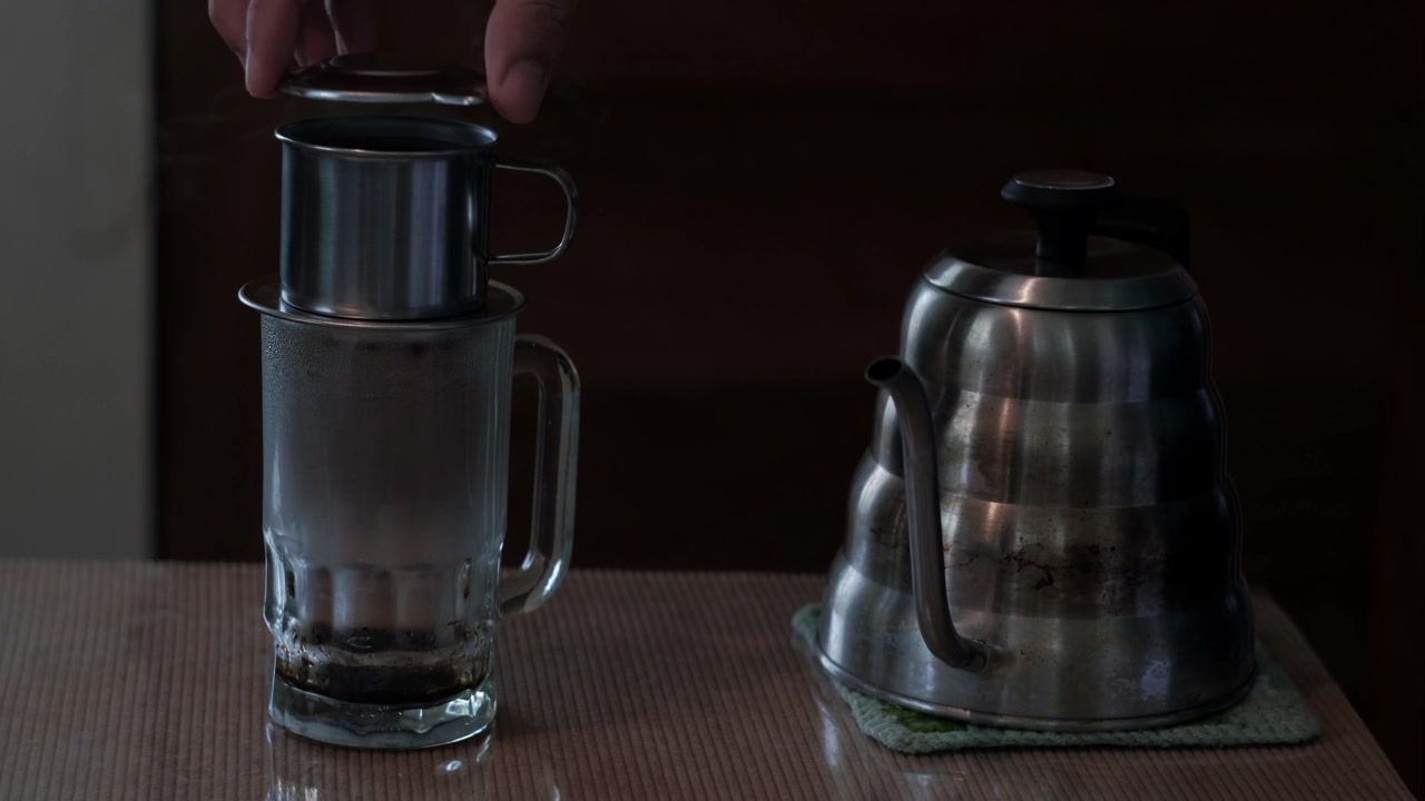 Statische Mittelaufnahme von heißem Wasser, das aus einem Schwanenhalskessel in einen vietnamesischen Phin-Filter während der Kaffeezubereitung im Innenbereich gegossen wird.