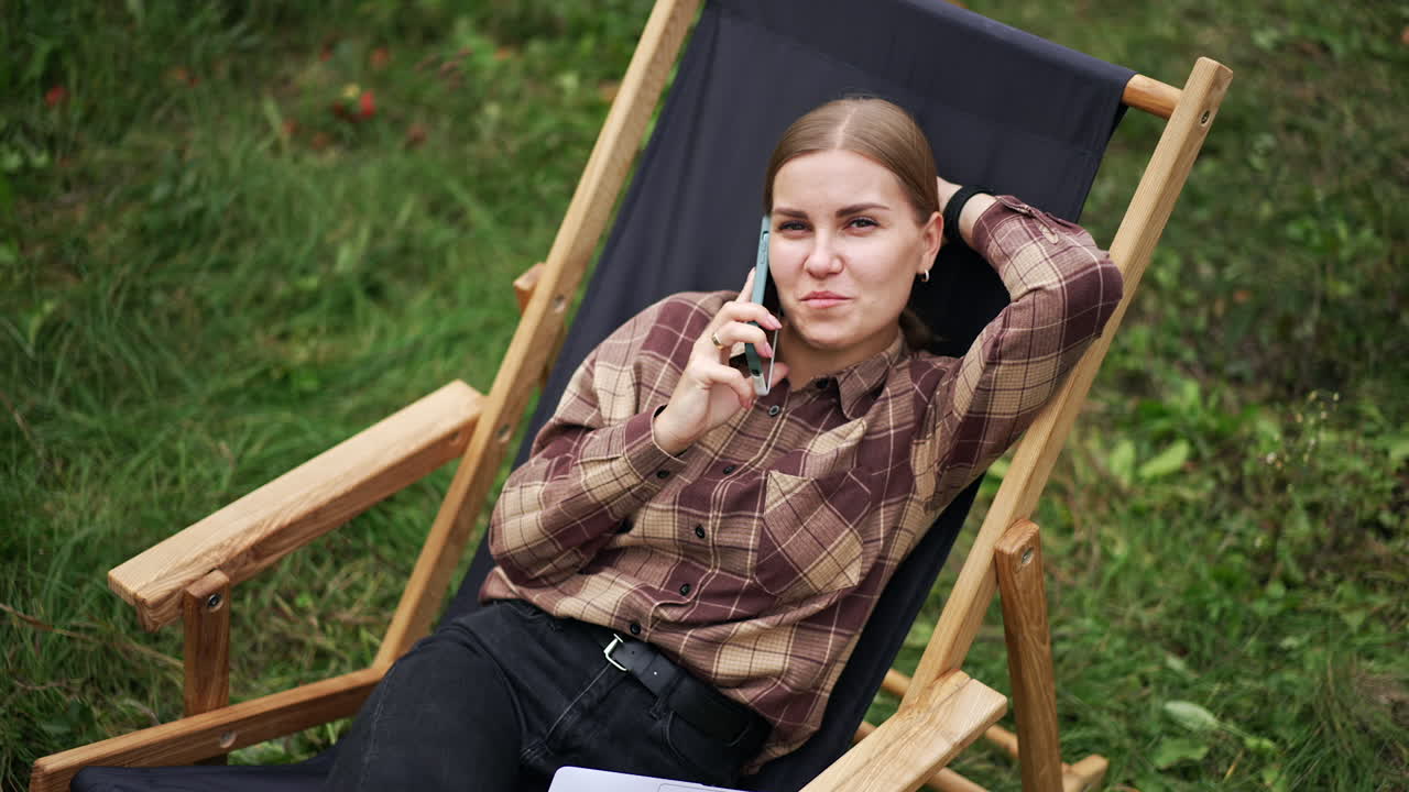 Relaxed happy Caucasian lady speaks on the phone lying in the garden chair. Free time freelancer having during remote work. Outdoors setting.