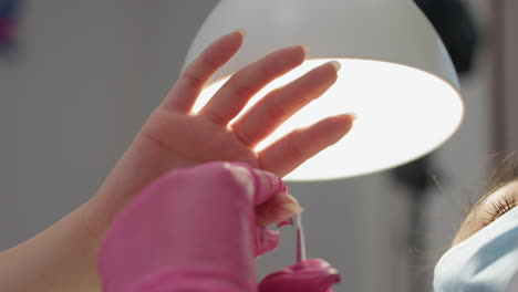 downward view of nail technician with visible mask and lashes wearing pink glove while carefully applying hardener along side of client thumb under strong white light during nail session