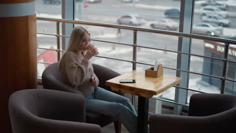 professional lady sipping coffee while sitting alone at wooden table in a mall with phone placed on the table, enjoying a break and observing the bustling city view outside