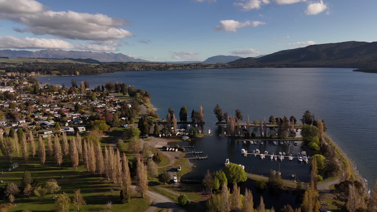 Aerial view of Te Anau Town as sunlight reflects across calm lake water near Te Anau Boating Club and boats. Marina with Fiordland mountains in background. Sunny autumn day in New Zealand