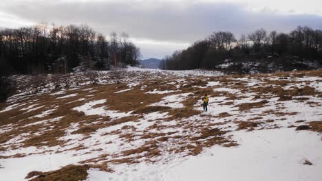 Wide-shot-of-a-hiker-walking-through-a-meadow-with-some-snow-and-a-forest-in-the-background