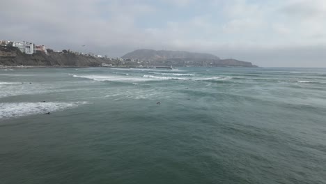 Langsame-Luftbildaufnahme-Vor-Der-Küste-In-Miraflores-In-Peru-Auf-Das-Türkisfarbene-Meer-Mit-Ruhigen-Wellen-Und-Surfern-Im-Wasser-Mit-Blick-Auf-Felsen,-Gebäude-Und-Berge-Im-Hintergrund