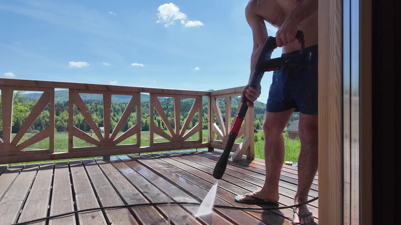 Shirtless man in blue shorts pressure washes a brown wooden terrace at a log cabin on a sunny summer day, surrounded by green meadows, forest and mountains.