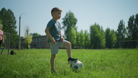 a young child places one foot on a soccer ball, looking focused, the background includes a partial view of his grandmother standing near a goal post