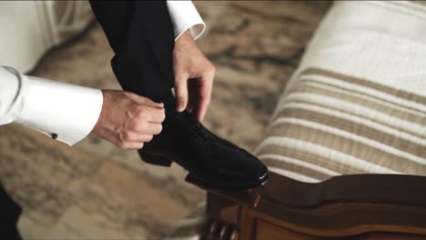 well-dressed man ties the shoelaces of his black leather shoes indoors, close-up view