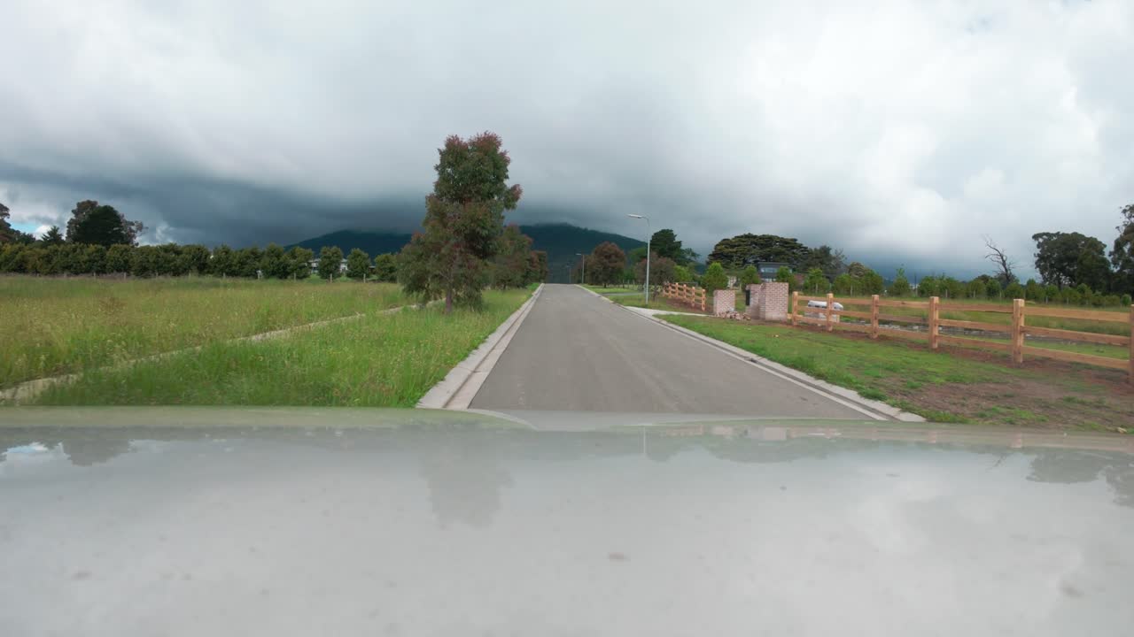 A scenic drive down a quiet road amidst lush fields and distant mountains. Cloudy skies hint at impending weather, creating a dramatic and serene atmosphere. Captivating wide shot