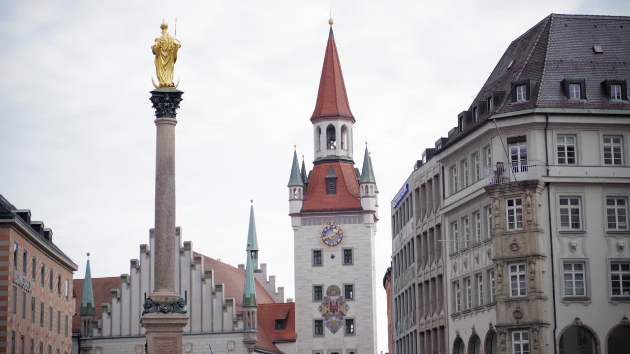The golden statue of Virgin Mary atop Mariensäule with Munich's Old Town Hall in the background. A historic cityscape at Marienplatz on a cloudy day, showcasing Gothic and Baroque architecture.