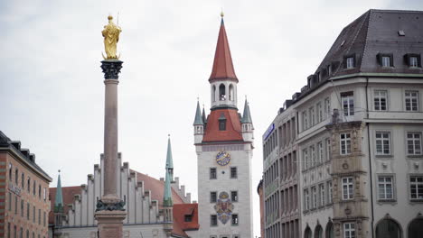 the golden statue of virgin mary atop mariensäule with munich's old town hall in the background. a historic cityscape at marienplatz on a cloudy day, showcasing gothic and baroque architecture.