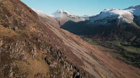 Vista-De-Retiro-Desde-Las-Montañas-Nevadas-Y-El-Valle-Verde-A-Lo-Largo-De-La-Ladera-De-La-Montaña,-Revelando-Un-Caminante-Sentado-En-El-Distrito-Del-Lago-Wasdale,-Reino-Unido