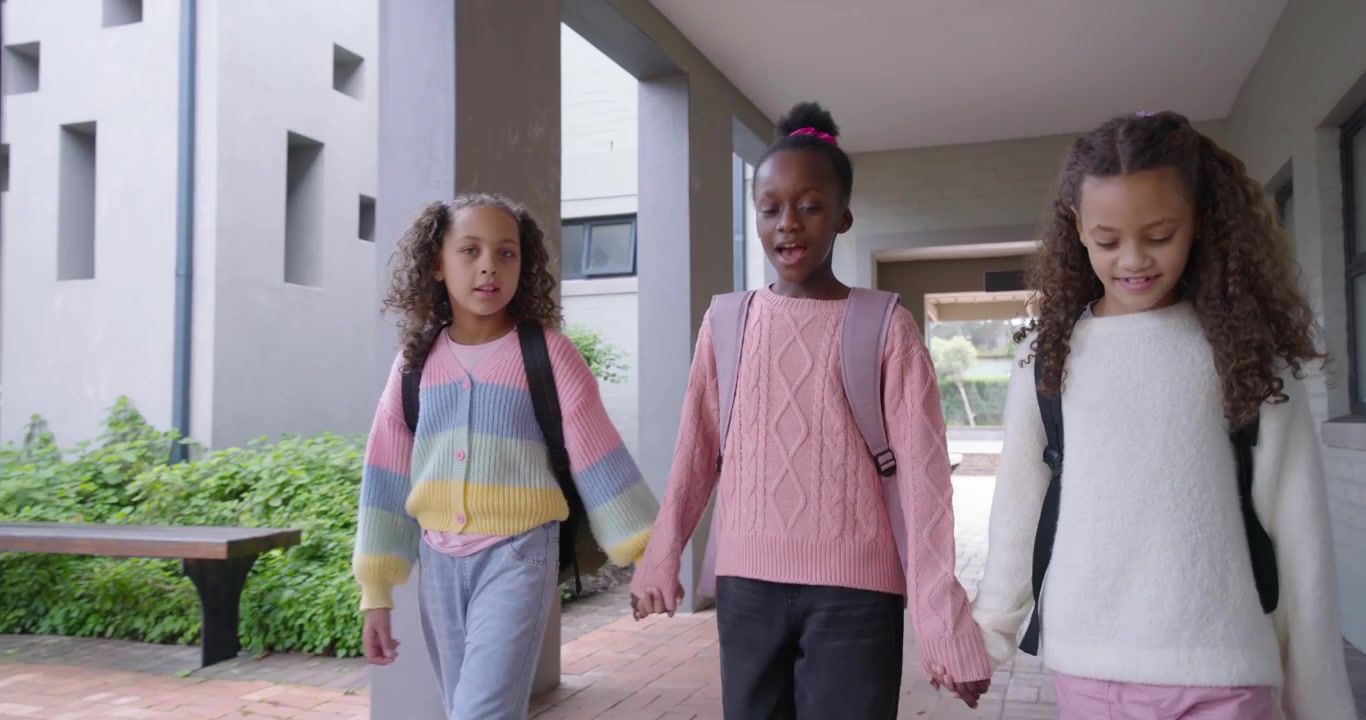 Diverse female preteens entering campus corridor holding hands walking to courtyard with backpacks