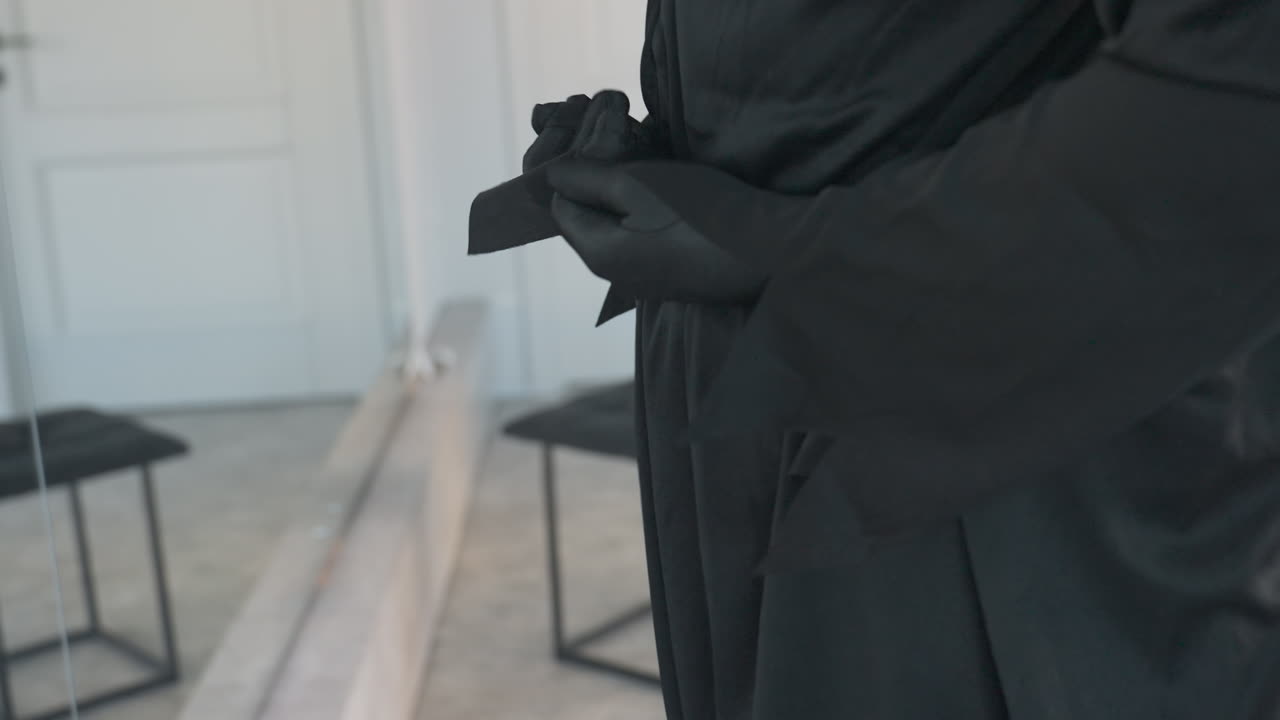 close up of person in flowing black costume tying rope belt around waist in front of floor to ceiling mirror, soft light highlights fabric folds and reflective surface