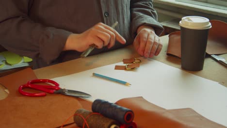 an experienced tailor in the workshop behind the workbench prepares accessories for creating a leather bag. glues together elements and drinks coffee during the break