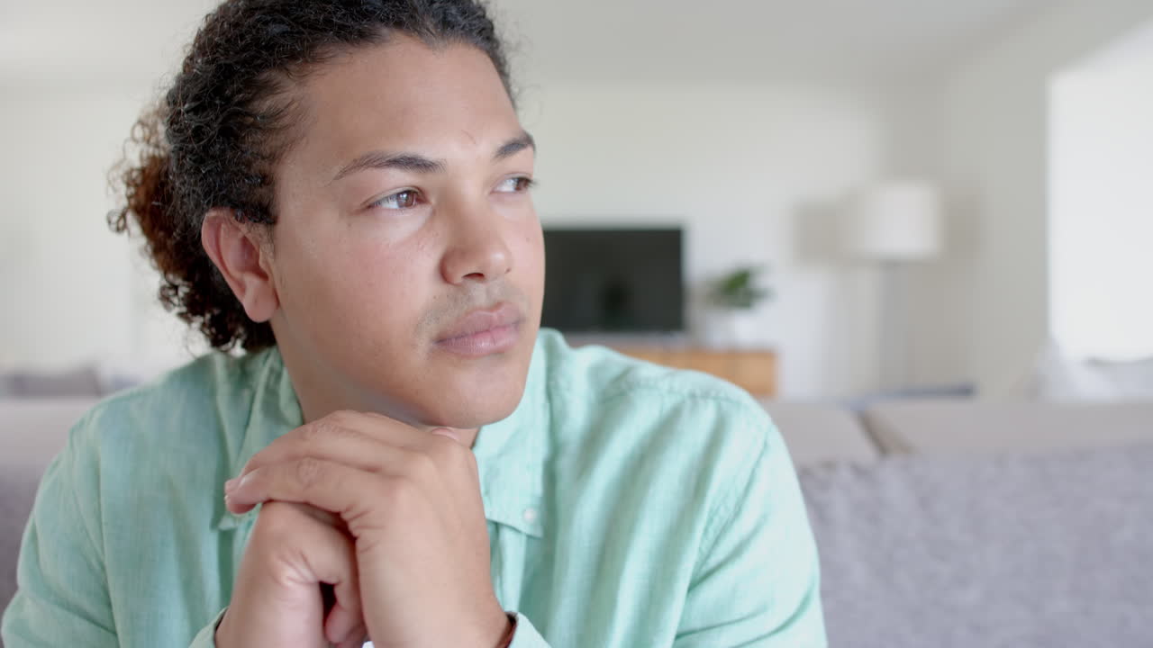 Thoughtful, sad biracial man with long hair sitting on sofa in living room, slow motion
