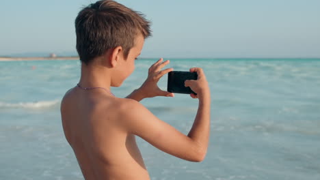 un chico tomando fotos con su celular en la playa, un chico filmando con su teléfono inteligente en la playa.