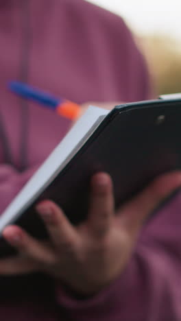 close-up of person in maroon top writing notes outdoors, holding clipboard with pen, with blurred background of sports field and court