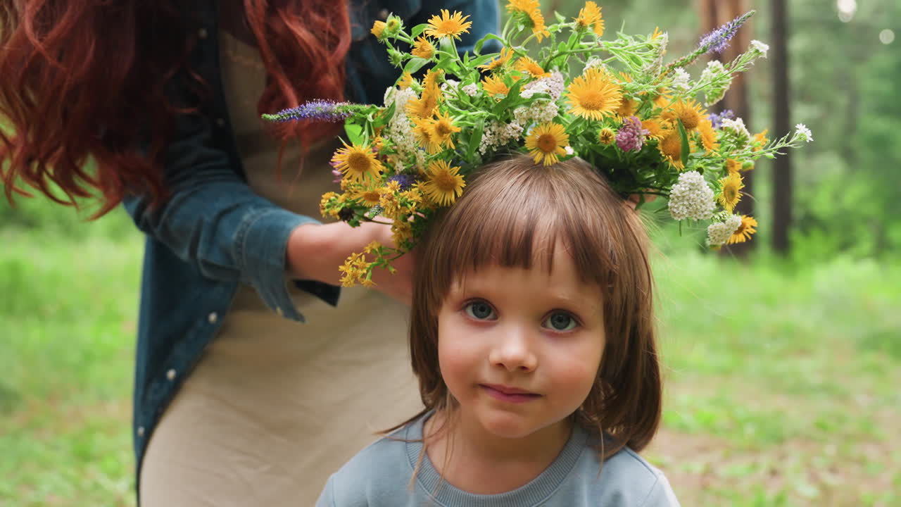 Portrait of little girl sitting outdoors while mother decorates her head with bright wildflower crown, surrounded by green forest, natural light, and warm summer atmosphere showing family love