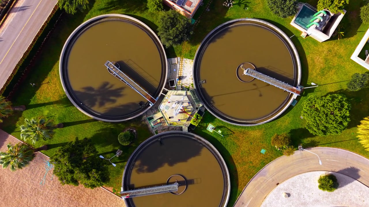 Overhead drone view of circular sedimentation tank at water treatment facility. Brown clarifier basin, solar panel roof, green lawn and palm tree shadow under bright daylight.