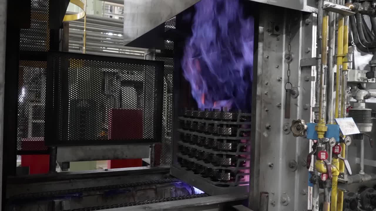 Automated conveyor transporting racks of metal gears into a high-temperature industrial furnace with vibrant blue and orange flames