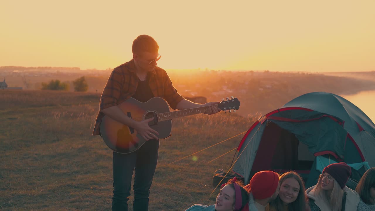 un ragazzo felice suona la chitarra agli amici seduti vicino alla tenda blu