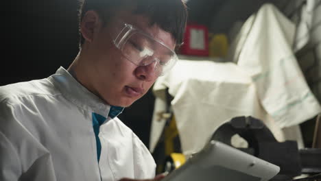 young chinese student in white lab coat focused intently on his tablet while wearing transparent safety glasses in an automotive workshop