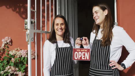 two smiling caucasian waitresses wearing aprons, standing in door, holding open sign