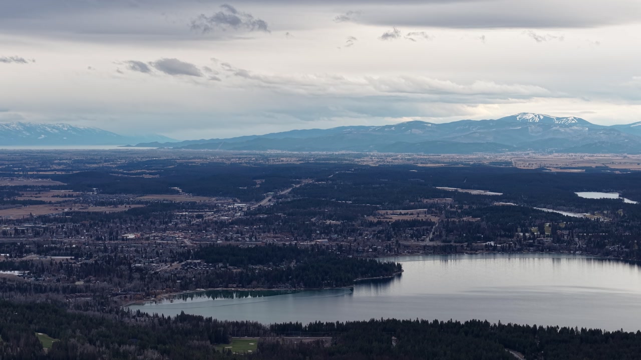 Aerial view of Flathead Lake with forested shoreline, distant snowcapped mountains and a cloudy sky in Montana.