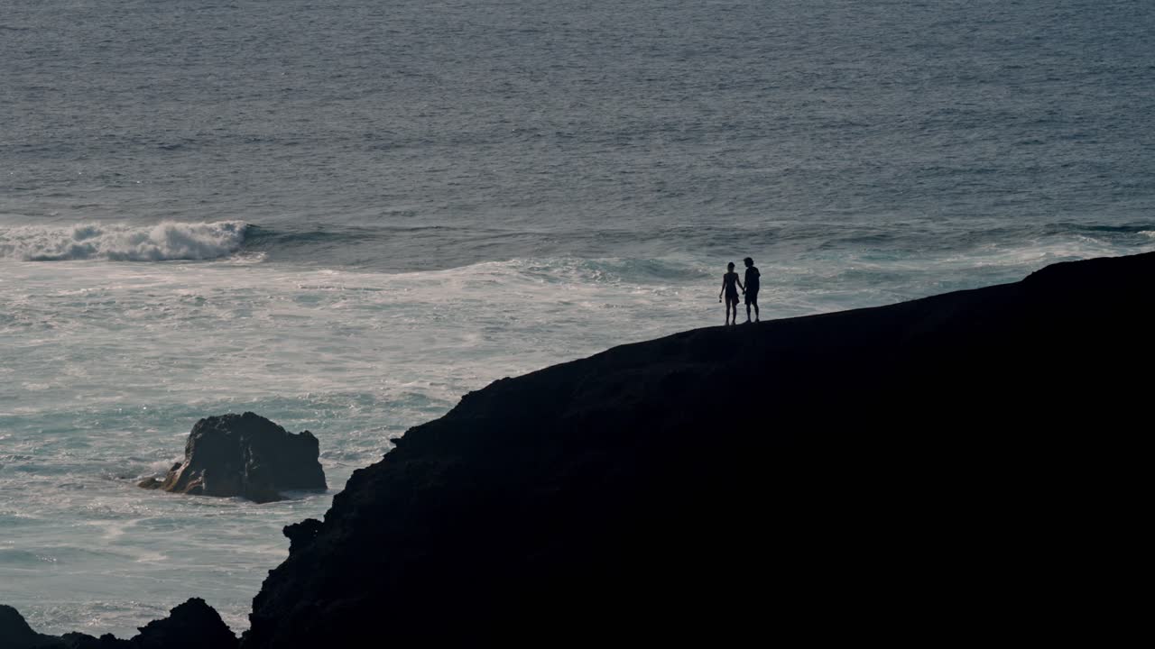 A young couple walks hand in hand along a rugged volcanic cliff overlooking the Atlantic Ocean in Lanzarote, Canary Islands, Spain.