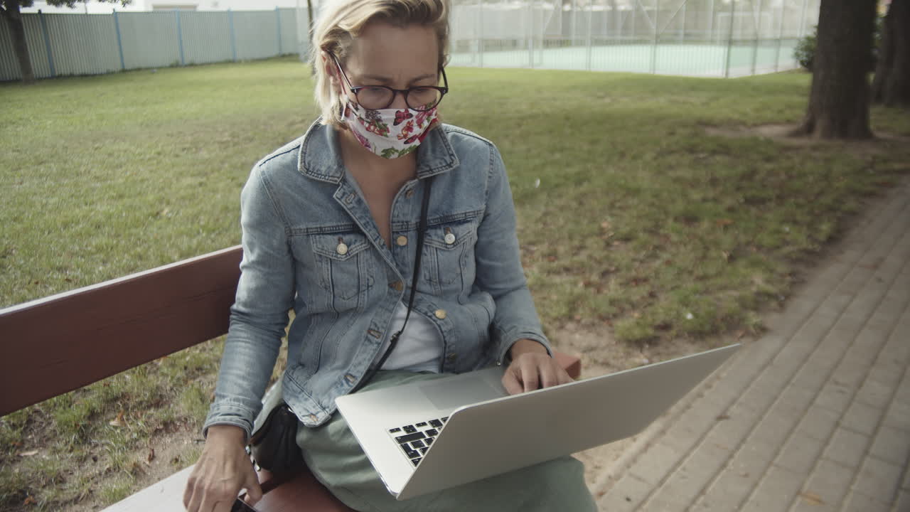 mujer con máscara y anteojos navegando en la computadora portátil y el teléfono inteligente mientras se sienta en el banco en el patio de recreo vacío - toma de cámara lenta media