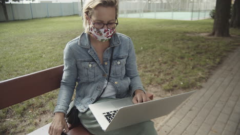 woman wearing mask and eyeglasses browsing on the laptop and smartphone while sitting at the bench in the empty playground - medium slowmo shot